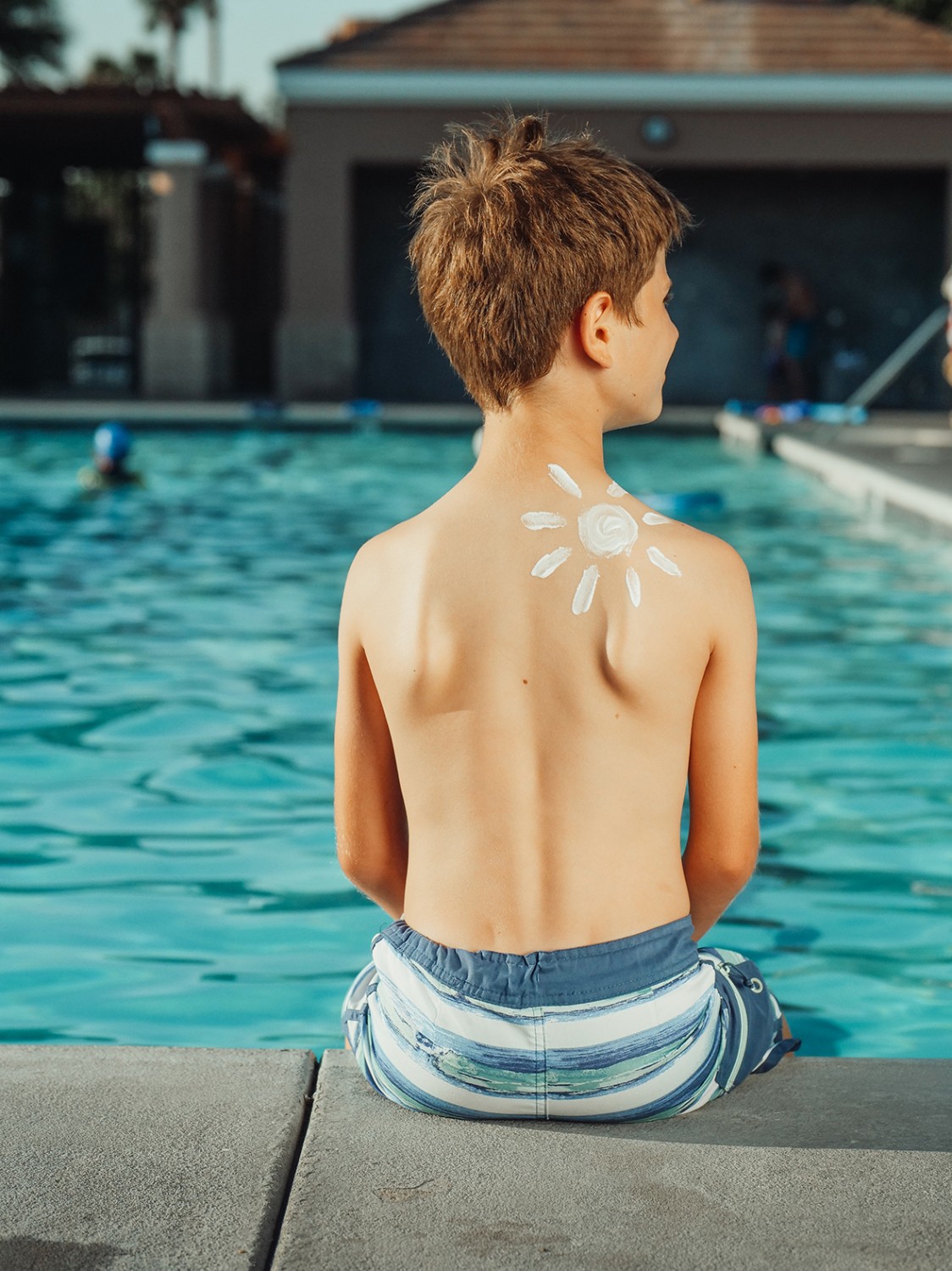 Chlapec sedí chrbtom pri bazéne a na pravom ramene má nakreslené slnko opaľovacím krémom.